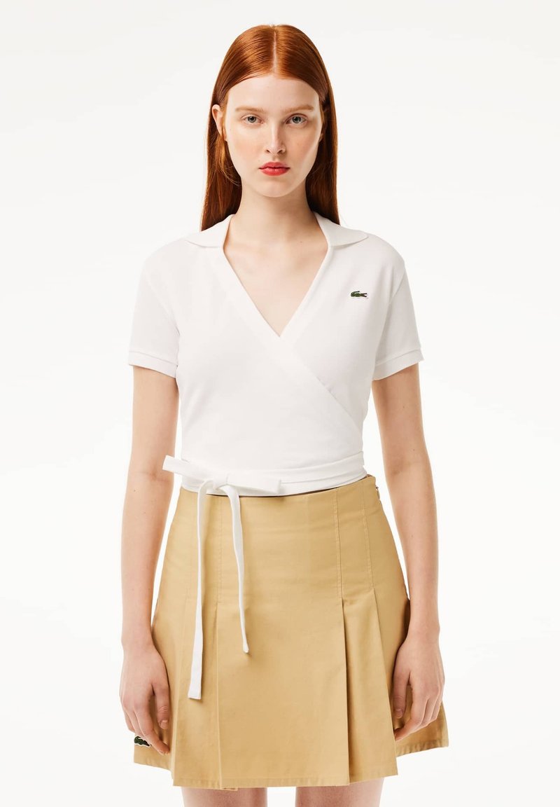 Top bianco avvolgente con scollo a V e maniche corte, abbinato a una gonna beige plissettata. Dettagli notevoli includono un nodo frontale e un logo verde.