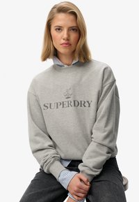 Szary sweatshirt z okrągłym dekoltem z grafiką "SUPERDRY", wykonany z miękkiej mieszanki bawełny, z opadającymi ramionami i ściągaczami, nałożony na niebieską koszulę w paski.
