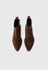 Bottines en suede marron avec un bout pointu et une fermeture éclair sur le côté. Elles présentent une semelle extérieure basse et texturée, ainsi qu'une languette à l'arrière.