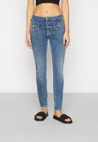 Mujer vistiendo jeans de mezclilla azul de talle alto con cierre de triple botón y sandalias negras de tejido, de pie contra un fondo sencillo.