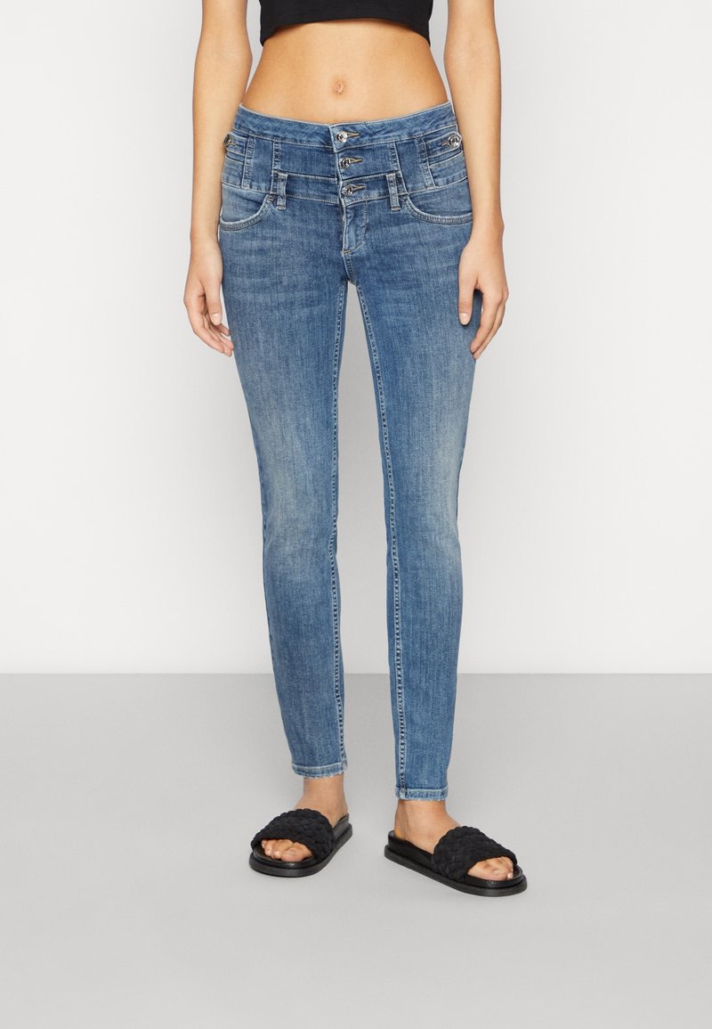 Mujer vistiendo jeans de mezclilla azul de talle alto con cierre de triple botón y sandalias negras de tejido, de pie contra un fondo sencillo.