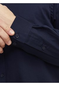 Laivastonsininen napillinen paita, jossa on teksturoitu kangas, hihassa kaksi nappia ja siisti muotoilu. Korosta hihan ja hihansuun yksityiskohtia.