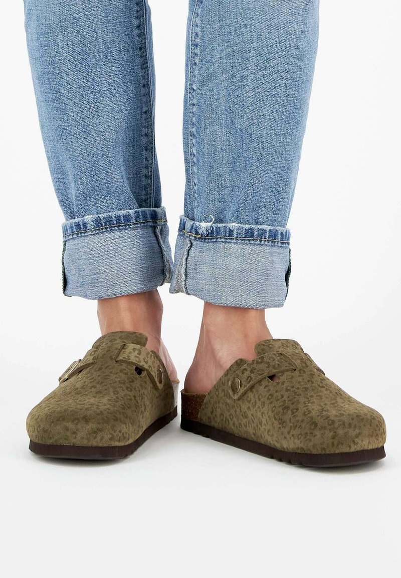 Grüne Leopardenmuster-Clogs aus Wildleder mit offenem Rücken und dekorativer Verschlussbeschlägen. Hochgekrempelte Jeans runden das Outfit ab.