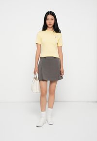 Gul kortermet t-skjorte, grå plissert miniskjørt med to svarte knapper, hvite sneakers og hvit håndveske, stående mot en ensfarget bakgrunn.