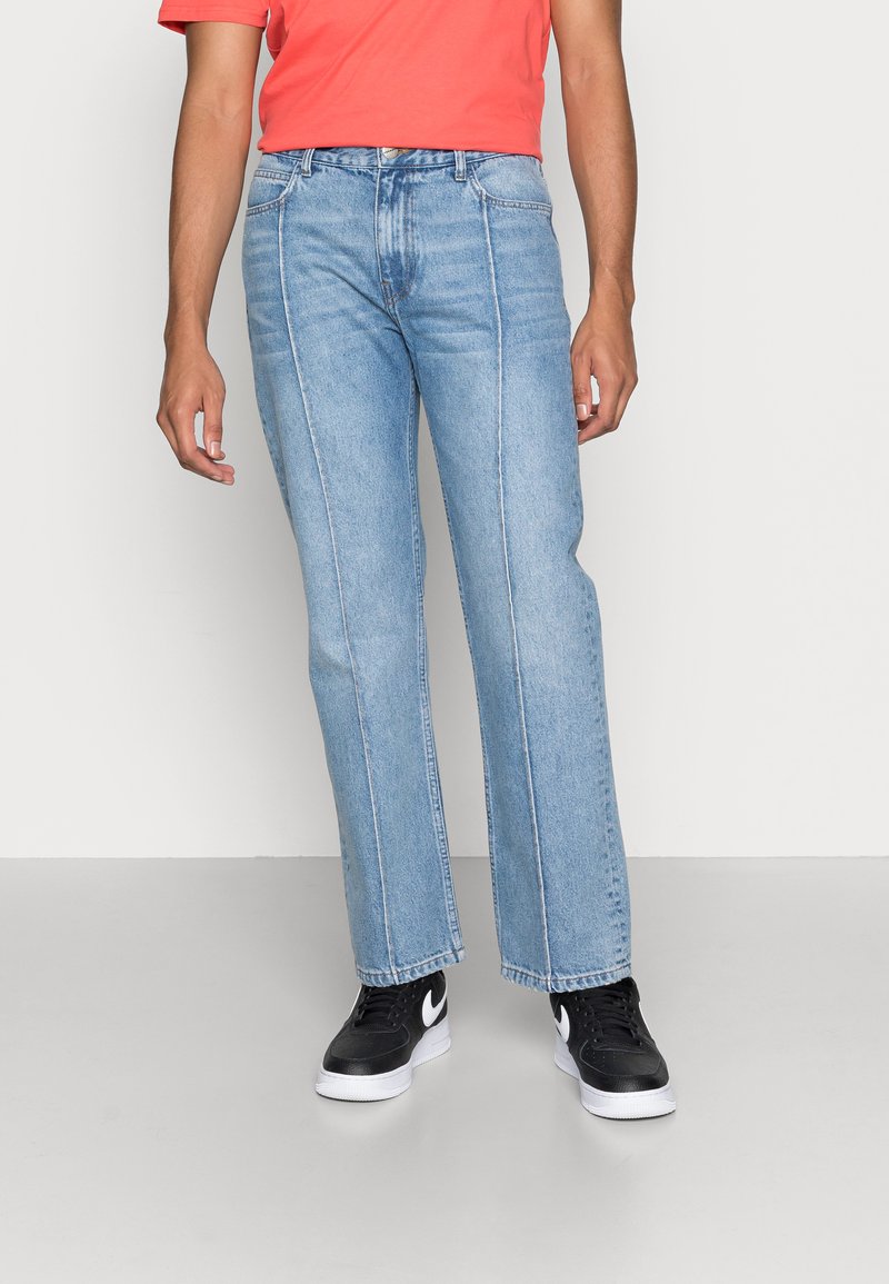 Jeans de ganga de corte reto em azul claro com costuras em contraste, usados com uma camisa vermelha. Ténis pretos com detalhes brancos completam o visual.