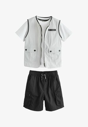 Balti trumpomis rankovėmis marškiniai su šviesiai pilka berankove liemene, kurioje yra priekinės kišenės, derinami su juodais kargo šortais, turinčiais kišenes su sagomis ir raišteliu juosmens srityje.