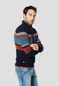 Pulover bleumarin cu model cu dungi multicolore în albastru, roșu și alb. Prezintă un guler înalt, tricot texturat și manșete și tiv striate.