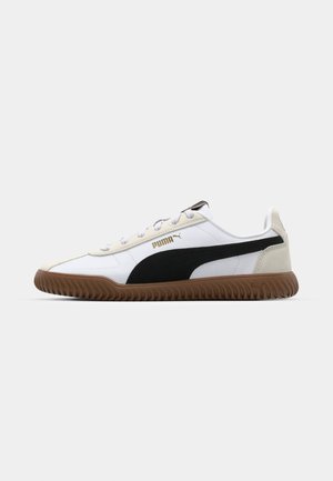 Scarpa sportiva bianca con accenti neri e beige, caratterizzata da una suola in gomma marrone texturizzata e un design con lacci. Logo dorato sul lato.