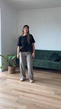 Čierne tričko s krátkym rukávom, béžové široké nohavice a biele tenisky; stojí v miestnosti so zelenou pohovkou a kvetináčom s rastlinou. Drevená podlaha.