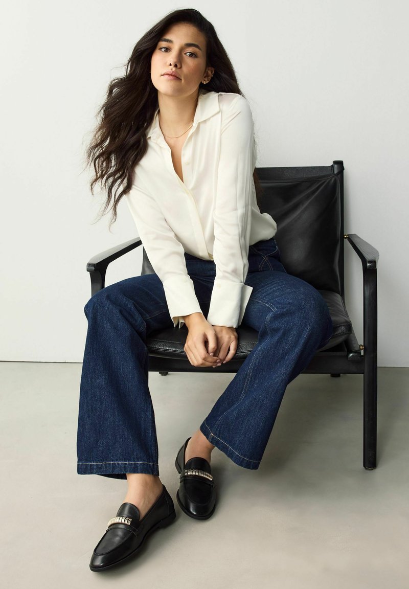 Camicia bianca con bottoni, jeans scampi in denim scuro e mocassini neri con un accento metallico. Donna seduta su una sedia nera.