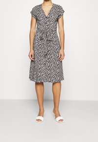 Donna che indossa un vestito nero fino al ginocchio con stampa floreale bianca, maniche corte, bottoni frontali e una cintura in vita, abbinato a sandali con tacco bianchi.