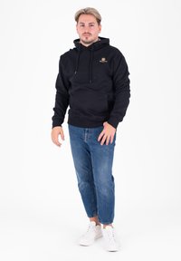 Felpa nera con cappuccio e tasca a marsupio, caratterizzata da un piccolo logo dorato. Abbinata a jeans azzurri chiari e sneakers bianche.