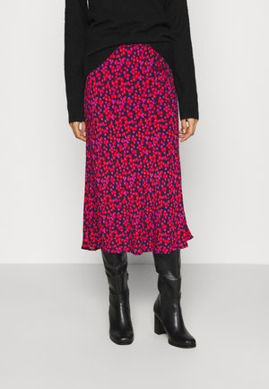 Femme portant un pull noir, une jupe violette avec motif floral rouge et rose, et des bottes noires hautes à talons, debout sur fond blanc.