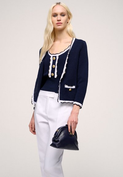 Námornícky blejzer s bielym lemovaním a zlatými gombíkmi, v kombinácii s ladiacim tielkom. K outfitu patria biele tailored nohavice a malá námornícka kabelka.