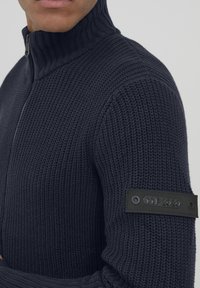 Maglione lavorato a maglia di un blu navy scuro con colletto alto, dotato di una zip frontale e una toppa nera rettangolare con testo in rilievo sulla manica.