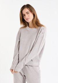 Felpa grigia chiaro a coste con maniche lunghe, scollo rotondo e dettagli con bottoni sulla spalla. Il tessuto sembra morbido e strutturato.