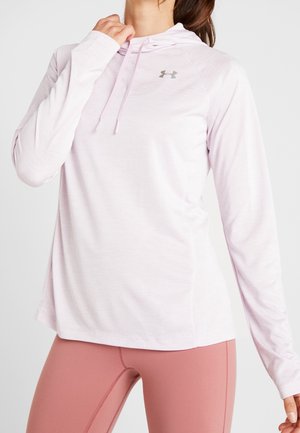 Frau trägt ein hellrosa langärmeliges Under Armour Hoodie und altrosafarbene Leggings, posiert mit einer Hand in der Nähe ihres Halses.