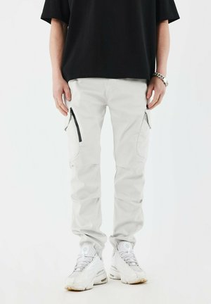 Pantalones cargo de color gris claro con dos bolsillos laterales con cremallera, piernas ajustadas y una textura suave, combinados con zapatillas deportivas blancas y una camiseta negra.
