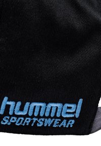 Nærbillede af sort stof med lyseblåt broderet "hummel SPORTSWEAR" logo på tekstureret materiale.