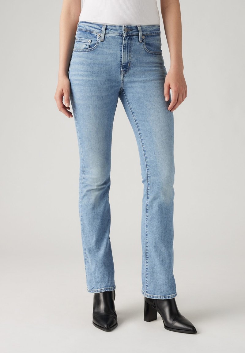 Femme portant un jean droit bleu clair et des bottines noires à talons sur un fond uni.