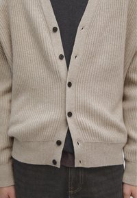 Beige geribde cardigan met een V-hals, voorzien van donkere knopen en een aansluitend ontwerp over een donker T-shirt, gecombineerd met donkere jeans.