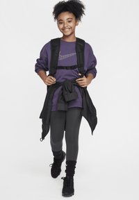 Sudadera morada con logo gris de Nike, mochila negra, chaqueta negra atada a la cintura, mallas grises y zapatillas negras con suelas gruesas.