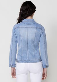 Világoskék farmerdzseki, testhezálló fazonnal, gombos gallérral és hátsó varrattal, két oldalsó zsebből és mandzsettagombokból áll.