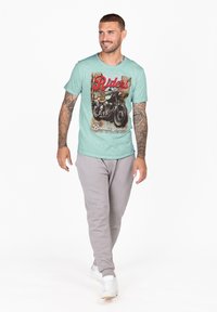 T-shirt verde menta con grafica 'Riders', abbinata a pantaloni della tuta grigi. La t-shirt presenta una stampa di motociclette vintage. Sneakers bianche indossate.