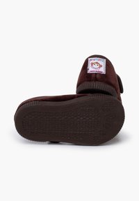 Braune Slipper zum Hineinschlüpfen mit einem weichen Stoff-Obermaterial, strukturierten Gummisohlen und einem bunten Affenetikett, das an der Seite genäht ist.