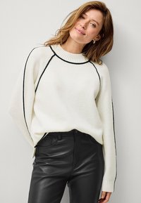 Pullover bianco lavorato a maglia con accenti neri lungo il collo e le cuciture, abbinato a pantaloni di pelle nera. Tessuto morbido e vestibilità rilassata.