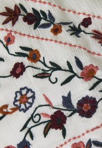 Tissu brodé avec un fond blanc présentant des motifs floraux multicolores en fils roses, orange, bleus et vert foncé, avec des détails complexes.