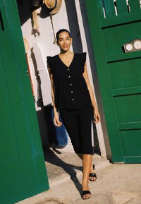 Femme en haut sans manches noir et pantalon capri sortant de portes doubles vertes avec des chapeaux accrochés à l'intérieur de l'encadrement.