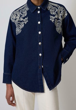 Donkerblauw denim overhemd met witte bloemrijke borduursels op de schouders, lange mouwen, zilveren knopen, nonchalant over ecru broek gedragen.