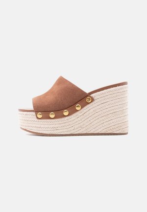 Sandalo zeppa in camoscio marrone con piattaforma intrecciata in corda, borchie dorate e un piccolo emblema dorato con marchio su sfondo bianco.