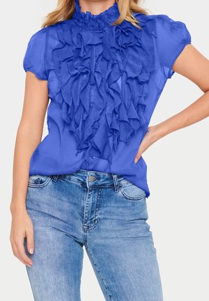 Frau trägt eine hellblaue, volantsbesetzte Bluse mit kurzen Ärmeln, kombiniert mit hellblauen Jeans, Hand in der Hüfte, einfarbiger Hintergrund.