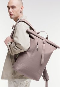 Zaino roll-top in un tenue mauve, realizzato in materiale resistente; presenta spallacci regolabili, una tasca frontale con zip e un logo in rilievo.
