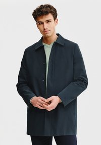 Ciemnogranatowy płaszcz z kołnierzem, przednimi guzikami i długimi rękawami, nałożony na jasnoniebieski sweter z okrągłym dekoltem. Gładka, matowa faktura.