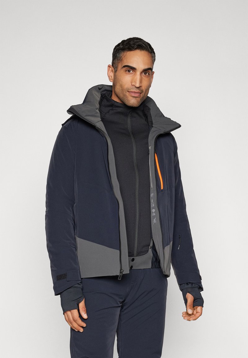 Giacca isolante navy e grigia con cappuccio, dotata di dettagli arancioni sulla zip e numerose tasche; texture morbida, design sportivo.