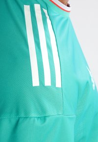 Turkos idrottströja med tre vita ränder på axeln, i kombination med en röd accent vid kragen och strukturdetaljer i tyget.