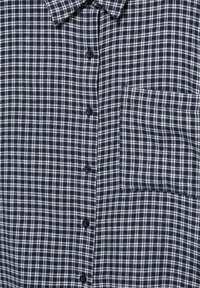 Camicia a quadri blu navy e bianca con colletto, chiusura frontale a bottoni e taschino sul lato sinistro del petto. Tessuto strutturato con motivo a griglia.