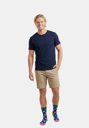 Homme souriant aux cheveux blonds portant un t-shirt bleu marine, un short beige et des chaussettes bleues avec des motifs colorés de fast-food, debout devant un fond blanc.