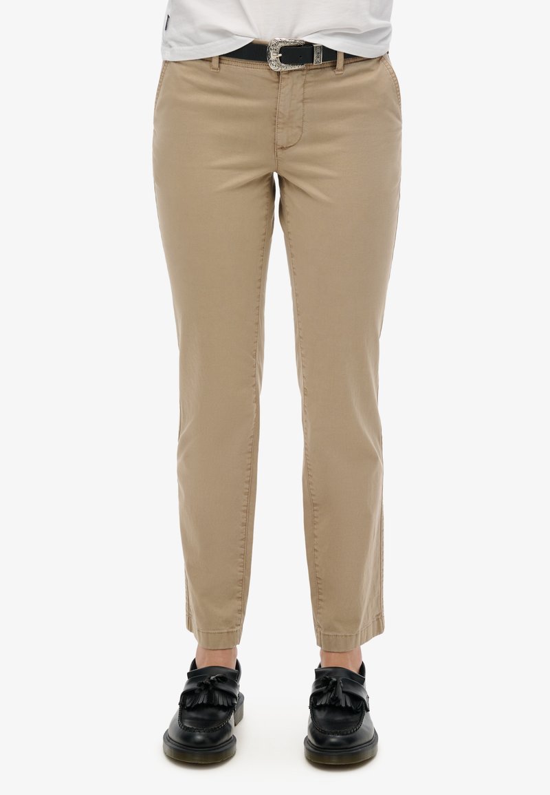 Chinos beiges avec une coupe slim, présentant une texture lisse, des poches avant et une ceinture noire avec une boucle décorative.