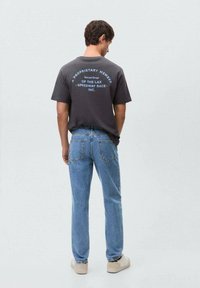 Gray t-shirt with blue graphic text on the back, paired with light blue jeans and beige sneakers. Plain background, standing posture.