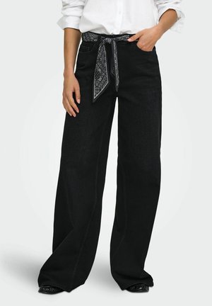 Femme portant un jean noir à jambes larges avec une ceinture en foulard à motifs, une chemise blanche aux manches retroussées et des chaussures noires.