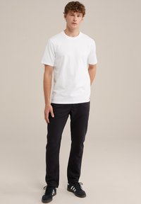 Vit bomullströja med korta ärmar, kombinerad med svarta slim-fit byxor och svarta sneakers med vita ränder. Neutral bakgrund.