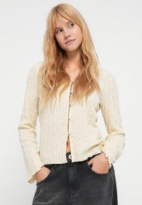 Cardigan crème texturé avec décolleté en V, manches longues et boutons décoratifs sur le devant. Associé à un jean foncé.