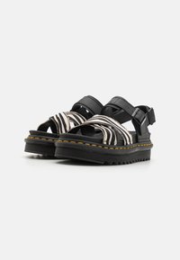 Sorte plattform sandaler med tykke såler, gul søm og zebra-stripete frontremmer, med justerbare sorte spenner ved anklene.