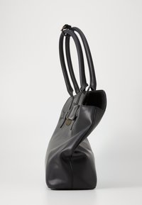 Sac tote en cuir noir avec des poignées épaisses et courbes, une forme structurée mais douce, et un détail en émail doré à l'avant.