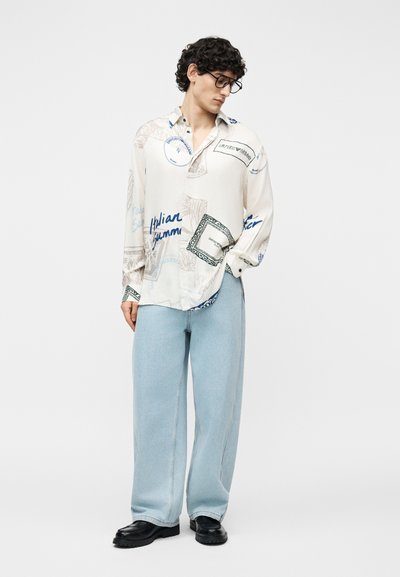 Homme aux cheveux bouclés foncés et lunettes portant une chemise blanche à manches longues imprimée, un jean large bleu clair et des chaussures noires.