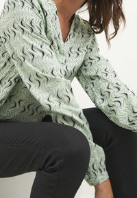 Vaaleanvihreä pusero mustalla aaltoilevalla kuviolla, v-kaula-aukolla ja pitkillä hihoilla, yhdistetty mustiin slim-fit-housuihin. Sileä rakenne, moderni muotoilu.